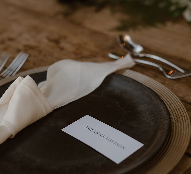 Black dinner plate with printed table name card, white napkins and silver cutlery on rustic wooden wedding table