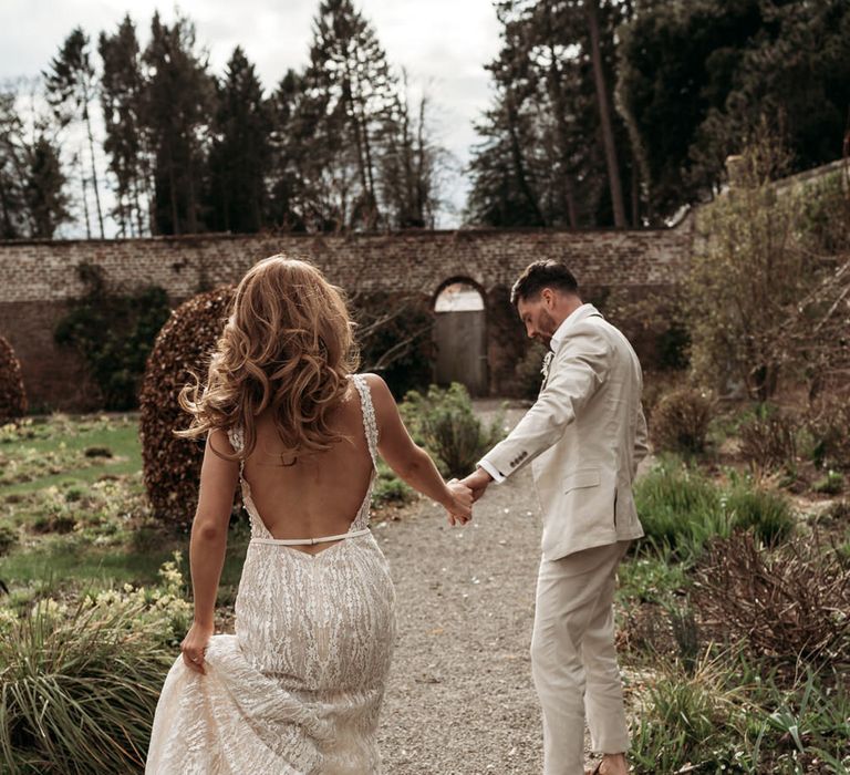 Bride in a low back wedding dress with long wavy hair holding her grooms hand in a beige suit 