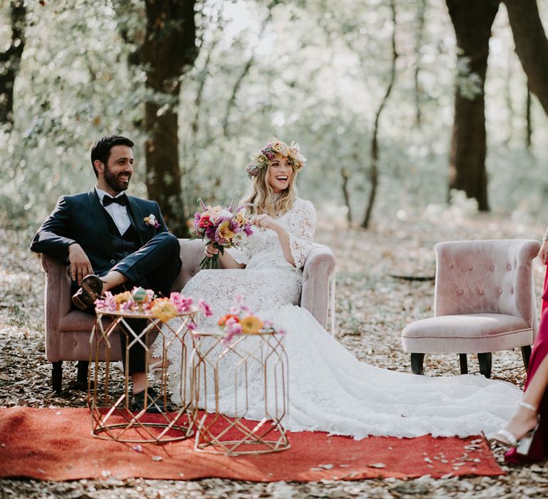 Bride in long sleeved lace boho dress and flower crown sits on pink velvet sofa with groom in navy suit in woodland with bridesmaids in long fuschia bridesmaids dresses at enchanted forest wedding in Italy