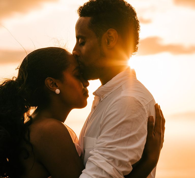 Groom kisses brides forehead as the sun sets behind them