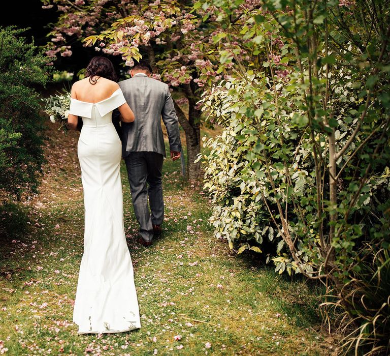 Bride in strapless Vagabond wedding dress with train walks into woods arm-in-arm with groom in grey Hugo Boss suit at lakeside wedding