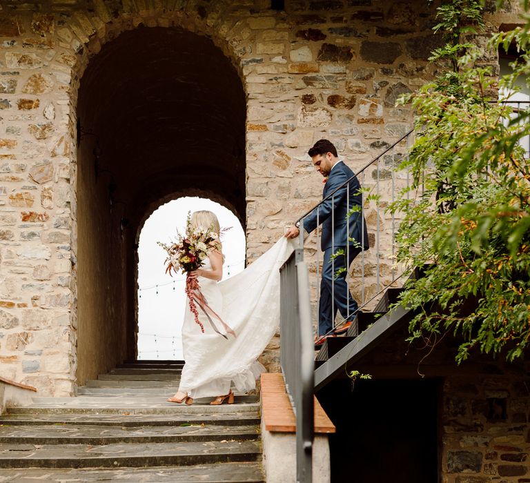 Bride and groom walking down a staircase, the groom is walking behind holding the bride's wedding dress above the ground