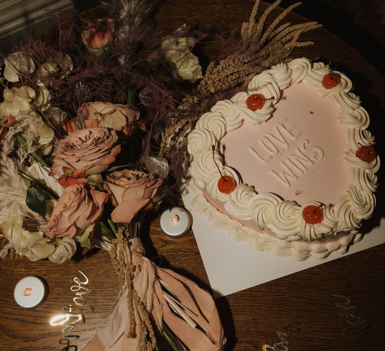 Heart shaped wedding cake with frosted 'Love Wins' next to bouquet