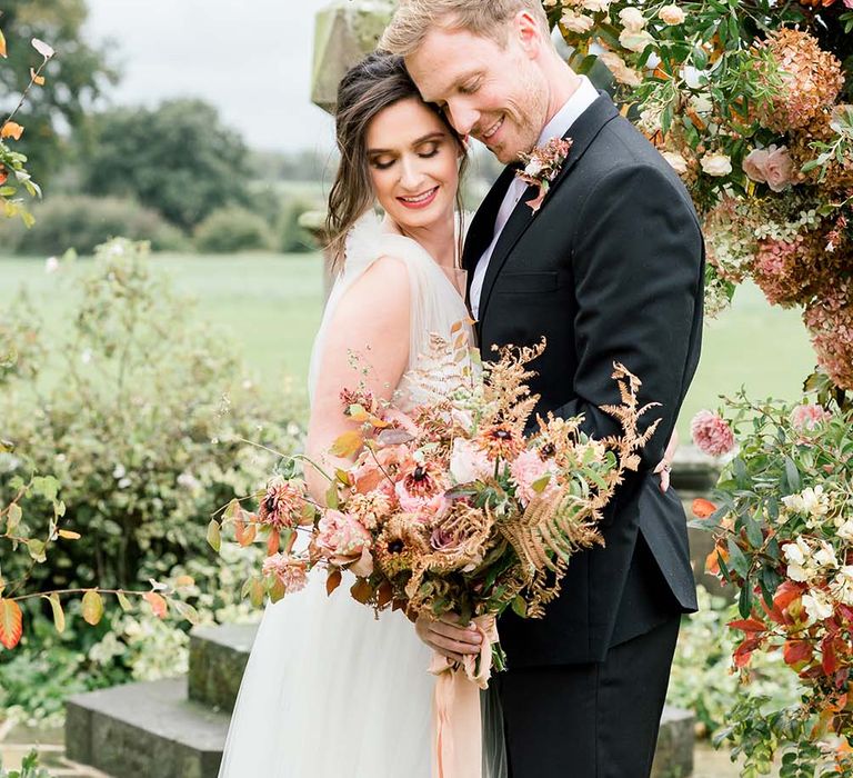 Bride and groom holding an autumn wedding bouquet together in from of a flower arch 