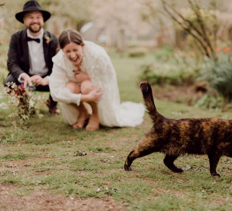 Laughing bride in white blouse, Charlie Brear wedding dress and applique veil holds multicoloured wedding bouquet watches brown cat whilst kneeling alongside groom in brown tweed suit, bow tie and black fedora at garden party wedding in Devon