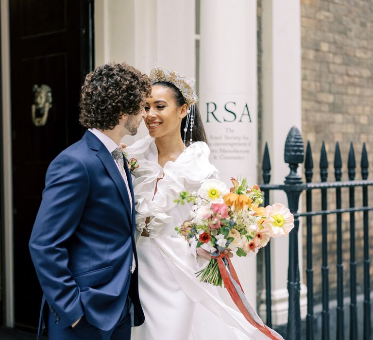 Bride & groom stand outdoors whilst looking lovingly at one another whilst bride holds floral bouquet with ruffled front