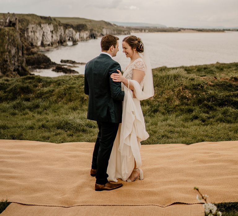 Groom in navy suit dances with bride in lace top capped sleeve wedding dress with satin skirt after Dunluce Castle wedding ceremony on clifftop