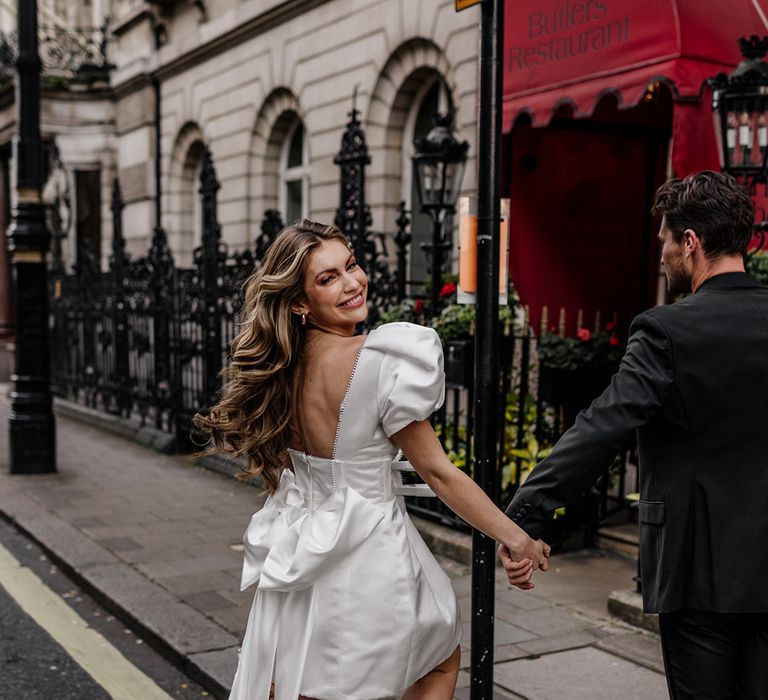 Bride with long wavy hair wearing a short wedding dress with bow back detail and pink mules, stepping onto the curb 