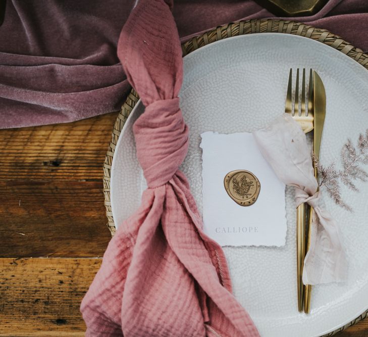 Place setting with wicker place mat, gold flatware, pink linen napkin, and gold wax seal name place card 