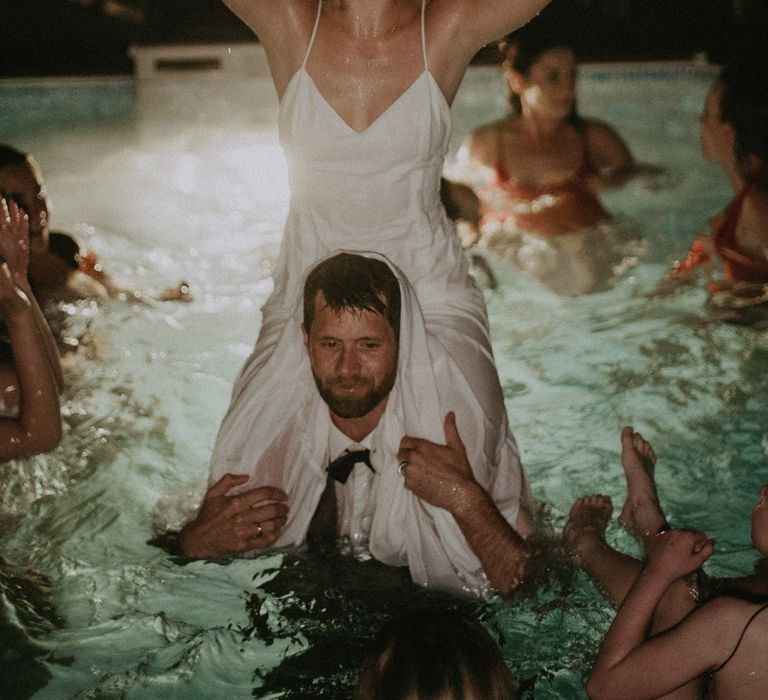 Bride in white Rime Arodaky wedding dress sits on grooms shoulders with arms in the air as they swim in pool at garden wedding with burnt orange wedding theme