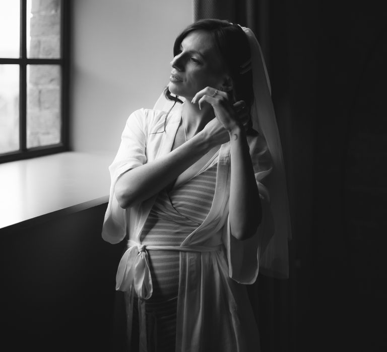 Bride looks out the window as she gets ready on the morning of her wedding whilst wearing white silk dressing gown