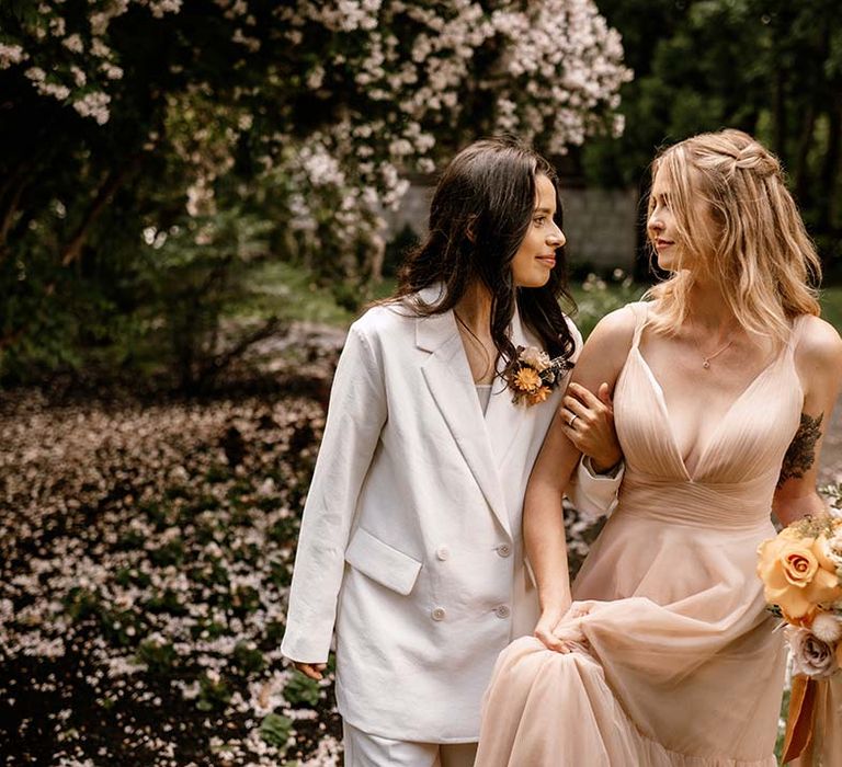 Bride in a white suit and a blush wedding dress holding a honey yellow wedding bouquet walking arm in arm 