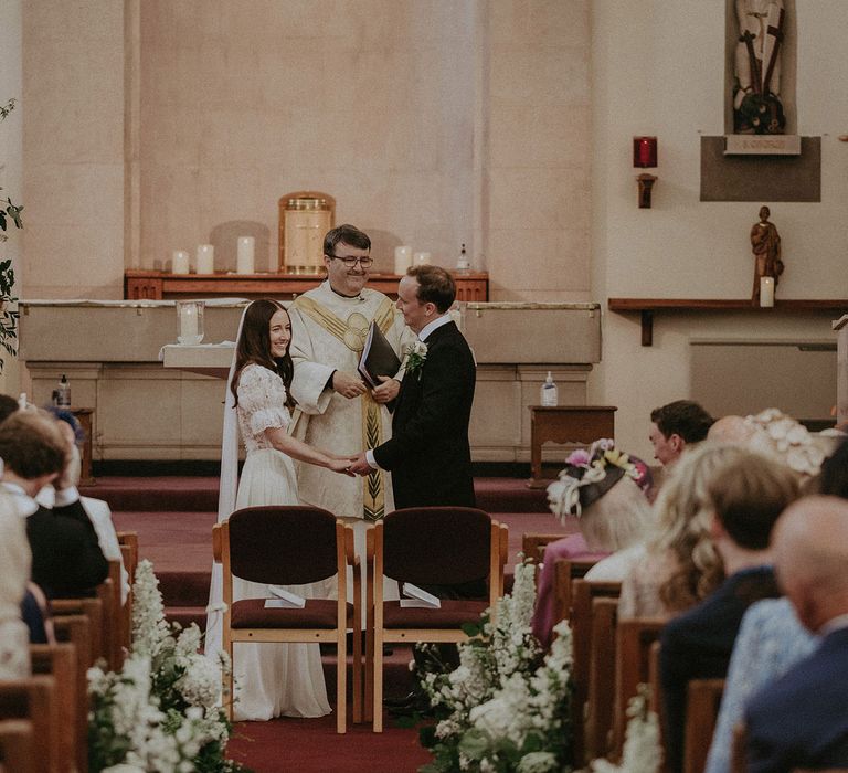 Bride in white lace puffed sleeve Daalarna wedding dress and bespoke veil smiles as she stands holding hands with groom in black morning coat at the alter during church wedding ceremony in Surrey