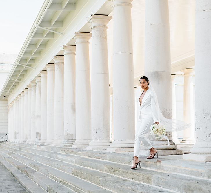 A bride wearing a white suit and veil walks down a set of stairs. There are white columns in the background behind her.