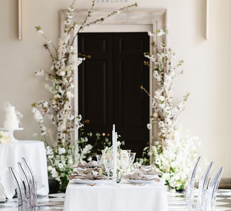 A table with 6 ghost chairs has a white table cloth, white candles and gold rimmed glassware for a black and white wedding reception. 