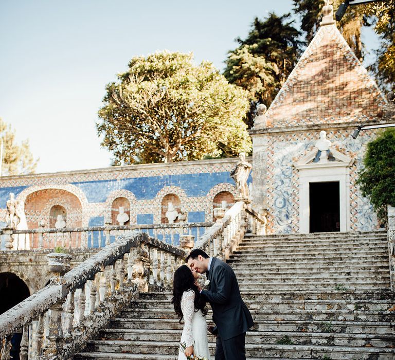 A couple kiss at outdoor wedding venue. They are stood on steps. He stands a few steps above and kisses her. Her long train is fanned out on the steps.