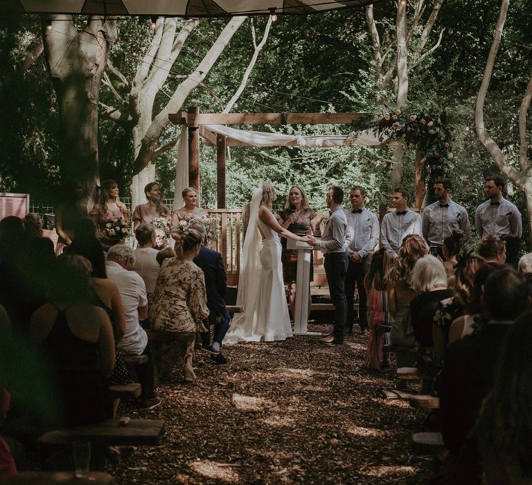 Bride & groom hold hands and look toward celebrant during their wedding ceremony outdoors in woodland setting