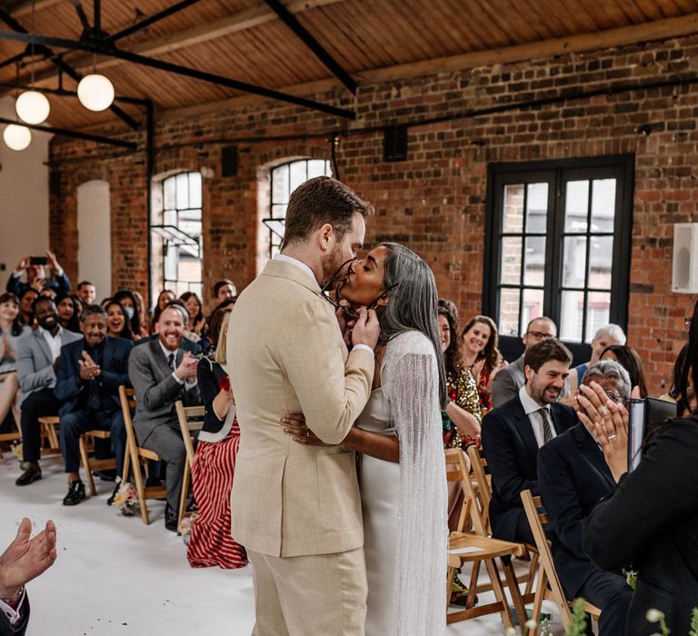 Groom in lane suit holding glasses kissed bride in satin Halfpenny London wedding dress and tasselled bridal cape during wedding ceremony at Loft Studios London