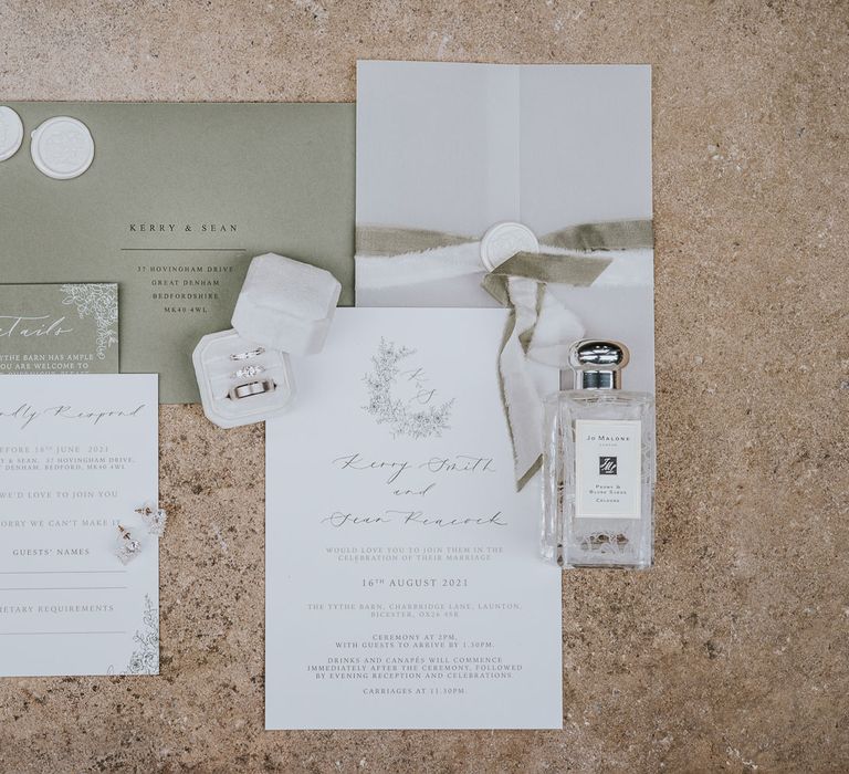 Green and white stationery with white wax seals, jo Malone perfume bottle, diamond earrings and wedding rings in white box at Tythe Barn wedding with barn wedding flowers