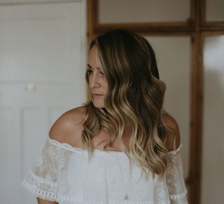 Bride with curled hair in Grace Loves Lace Bardot wedding dress and long silver necklace before Isle of Wight wedding with macrame wedding decor