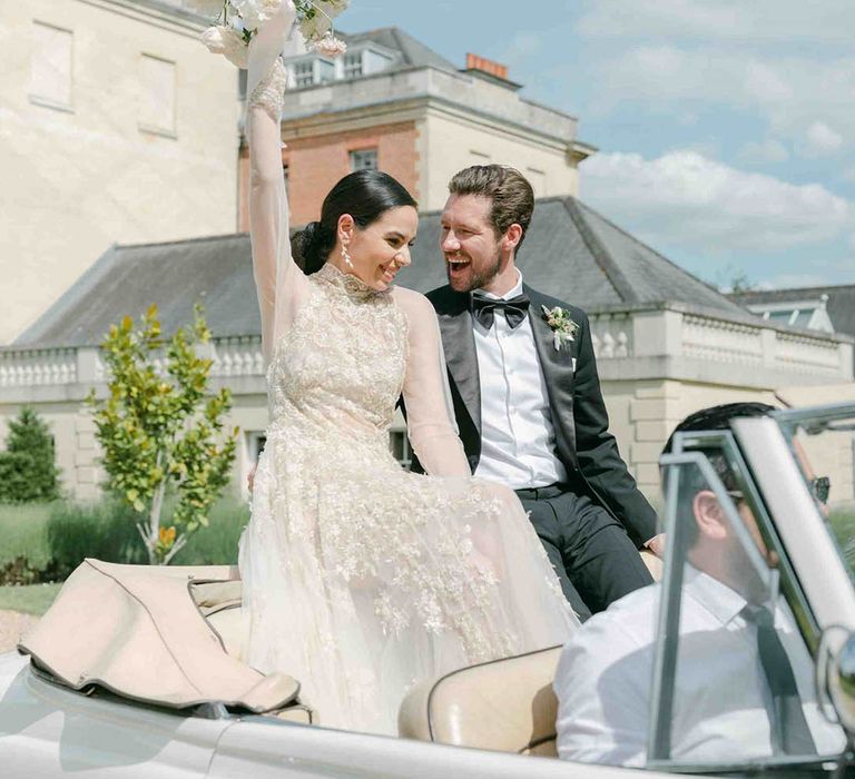 Bride in a lace long sleeve wedding dress sitting on the back of a vintage wedding car waving her bouquet int he air 