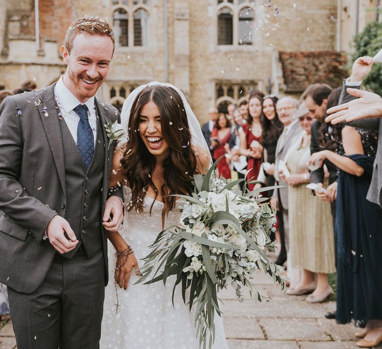 Laughing bride in Pronovias wedding dress and veil holding white and green bridal bouquet walks with groom in grey three piece shot, blue tie and white rose buttonhole as guests throw confetti at Notley Abbey wedding