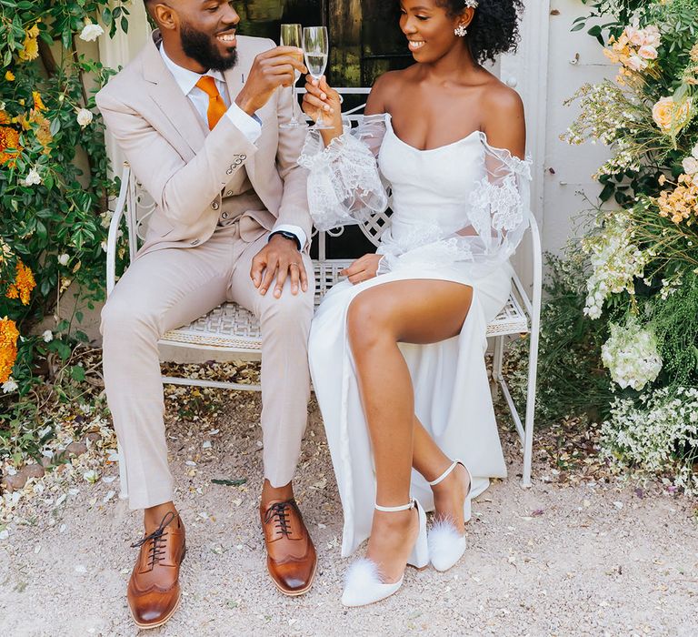 Bride in a strapless wedding dress with sheer sleeves drinking champagne with her groom in a stone coloured suit outside their Modern Hall wedding venue 