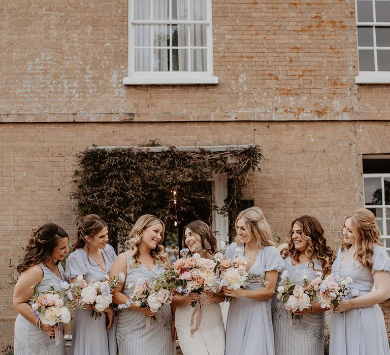 Bridesmaids wear lilac dresses in differing styles whilst holding pastel bouquets and looking toward bride