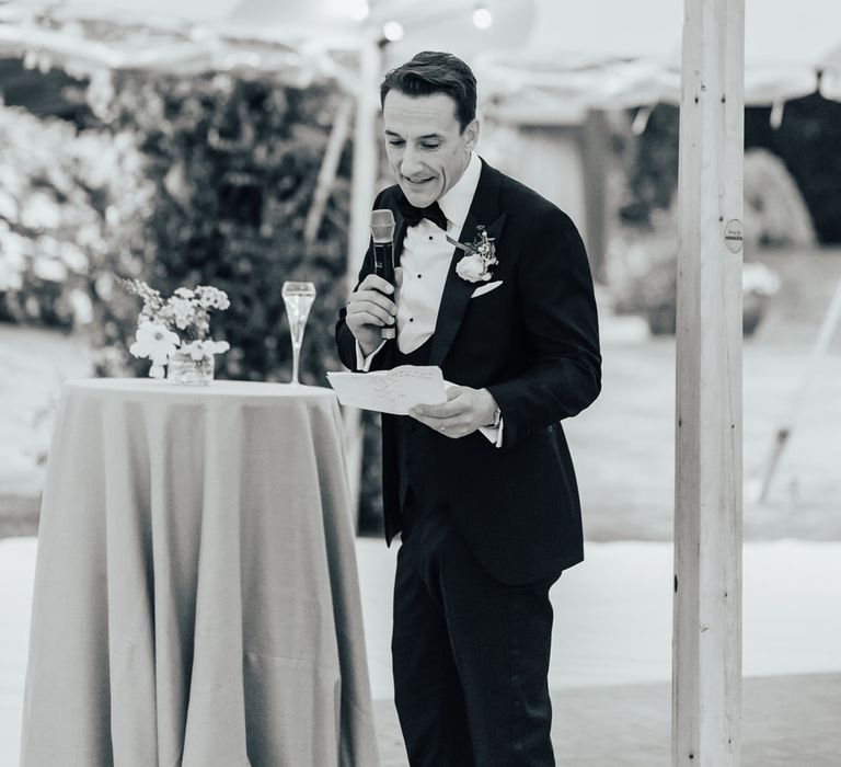 Groom in black tie with floral buttonhole holds mic as he makes wedding speech during sperry tent wedding reception in Hertfordshire