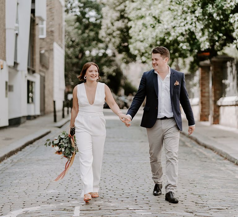 Groom in stone chinos, white shirt and blue Lazer holding hands with his bride in a jumpsuit at their London wedding 