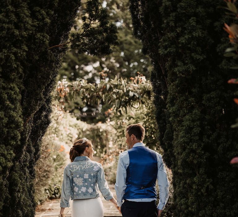 Bride & groom walk hand in hand and bride wears hand stitched denim jacket