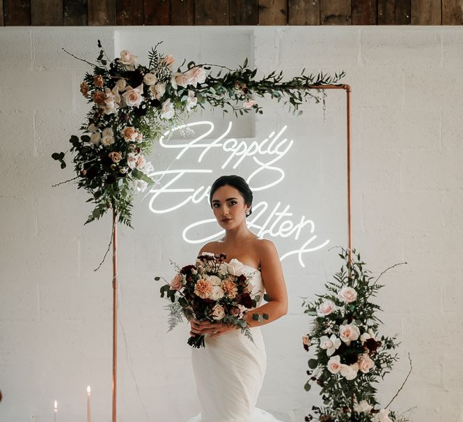 Bride stands in front of neon light and metal archway complete with floral decor