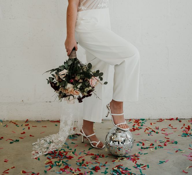 Bride wears jumpsuit complete with sheer top and embroidery whilst holding floral bouquet