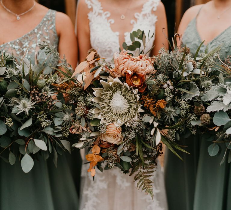 Bride in lace wedding dress and bridesmaids in sage green bridesmaid dresses hold green mixed thistle bouquets