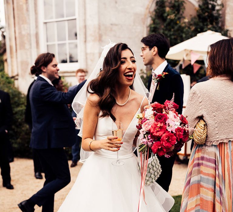 Bride in layered tulle skirt wedding dress with plunging neckline, pearl necklace and veil stands in garden holding pink, white and red bridal bouquet and glass of sparkling wine