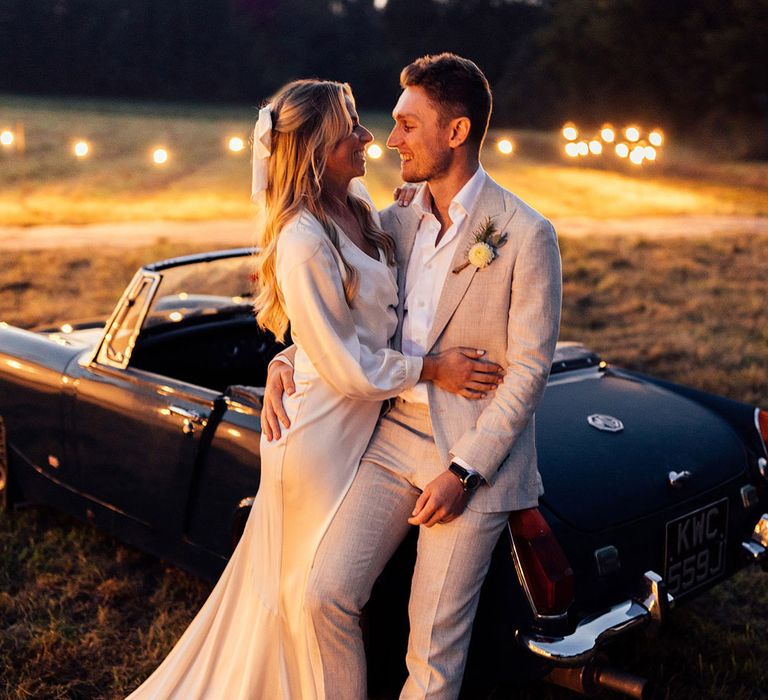 Bride in satin wedding dress with train and hair bow stands with groom in grey suit leaning on vintage convertible in field 