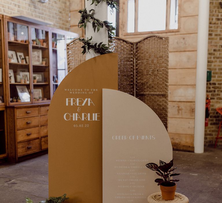 Wedding welcome sign and order of events notice board in mustard yellow and cream with potted plants and wicker details 