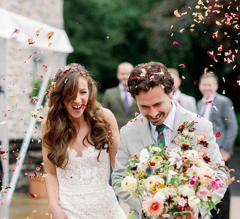 Bride in lace wedding dress with curled brown hair walks through confetti with groom in bespoke grey suit with green tie