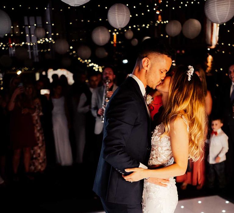 Bride and groom have their first dance and share a kiss on white dance floor with white lantern decorations in the background 