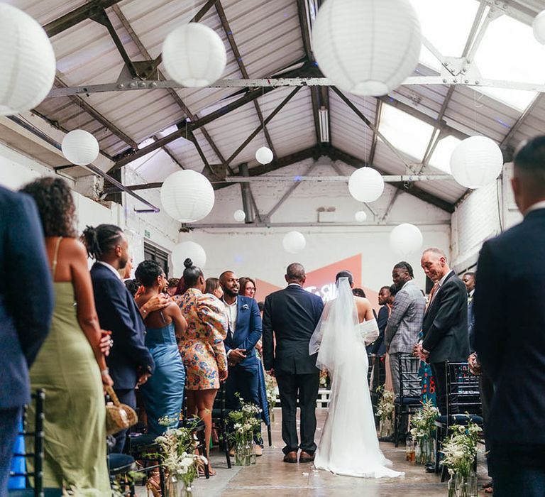 Bride is walked up the aisle by her father in black tie at industrial wedding venue