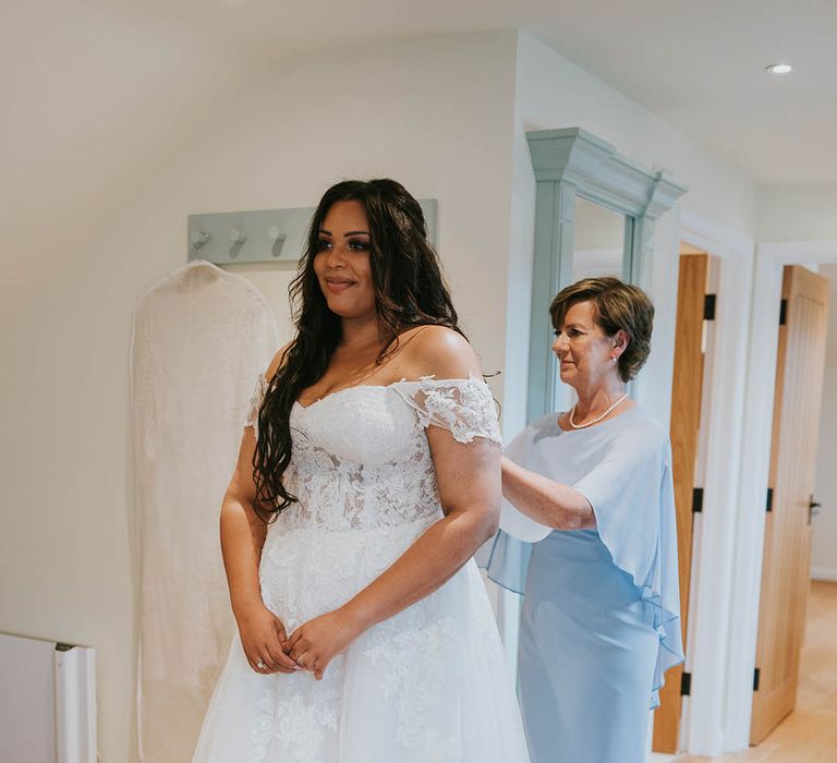 Wedding guest in light blue dress and pearl necklace helps the bride into her lace off the shoulder wedding dress