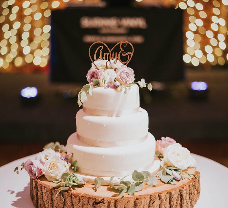 Short three-tier wedding cake with white icing, pearl decor and pale pink ribbon and pink and white roses with personalised wooden cake topper on tree trunk stand 