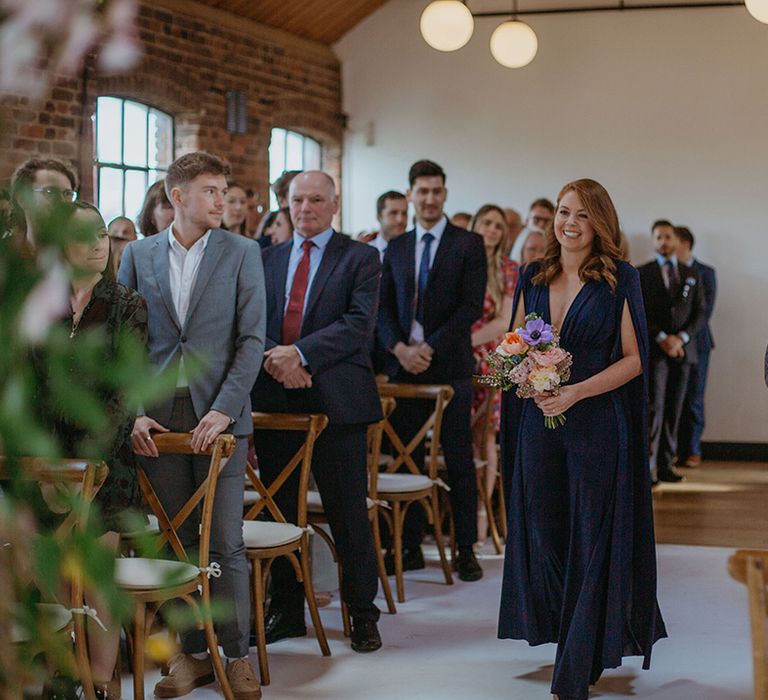 Bridesmaid in navy jumpsuit with cape arm detail walks down the aisle holding spring bouquet at Loft Studios 