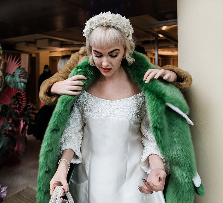 Bride with white flower headband and pearl handbag gets help putting on her green fur coat 