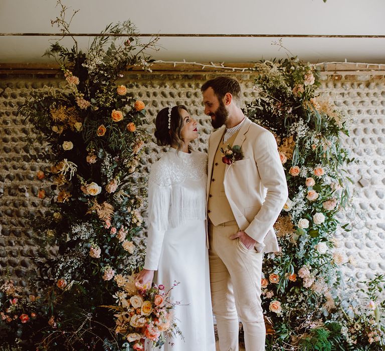 Bride in a long sleeve satin wedding dress with tassel bodice detail standing at the altar with her groom in a beige suit at Montague Farm Hankham