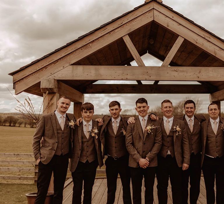 Groom with groomsmen in matching three piece brown suits posing outside at Eden Barn