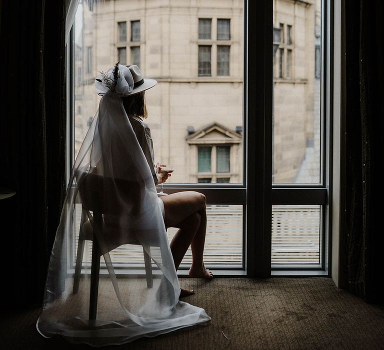 Bride wearing fedora hat with veil attached looking out of the window 