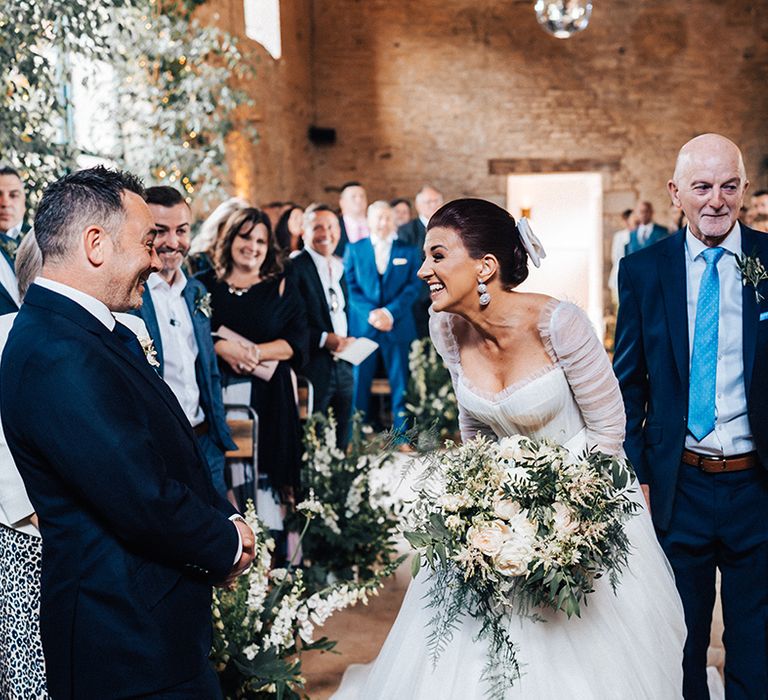 Bride with large white and green bouquet greets the groom in blue suit at the end of the aisle in ruffle wedding dress 