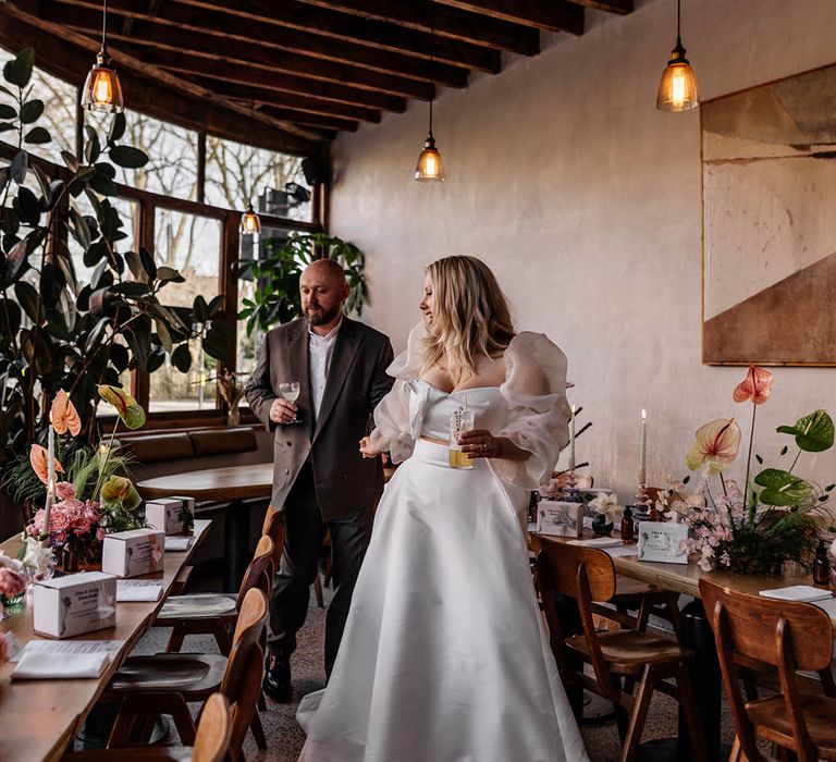 Bride in a Jesus peiro wedding dress holding hands with her groom in a double breasted jacket looking at their wedding reception table decor, flowers and favours 