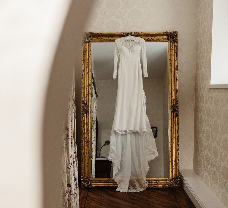 Lace bridal gown with long sleeves hangs in front of gold mirror 